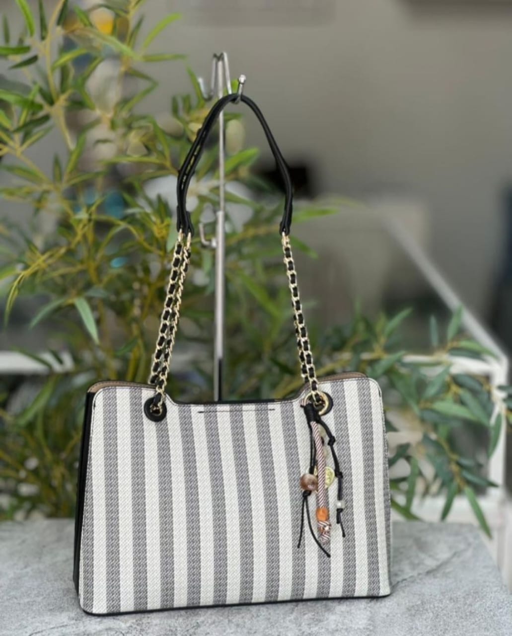 Vertical Striped Tote Bag is a casual everyday handbag featuring grey-and-white vertical stripes, sturdy leather-accented shoulder straps, and a decorative beaded tassel. Its structured, roomy design easily handles daily essentials for work or travel